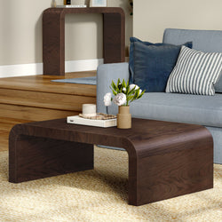 Modern dark walnut curved coffee table on jute rug with ceramic vase of white peonies, stacked books and candle beside a light blue sofa with navy and striped throw pillows in a neutral contemporary living room