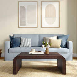 Modern living room with light blue sofa, curved dark wood coffee table, striped and textured throw pillows, abstract framed wall art and woven area rug