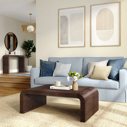 Modern living room with light blue sofa, striped navy pillows, curved dark wood coffee table with vase, jute rug and abstract framed wall art