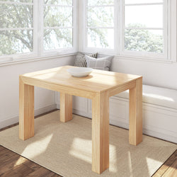 Sunlit light-wood dining table in a cozy breakfast nook with built-in white bench, gray check and striped pillows, white ceramic bowl, natural jute rug, hardwood floor and large white-framed windows overlooking trees