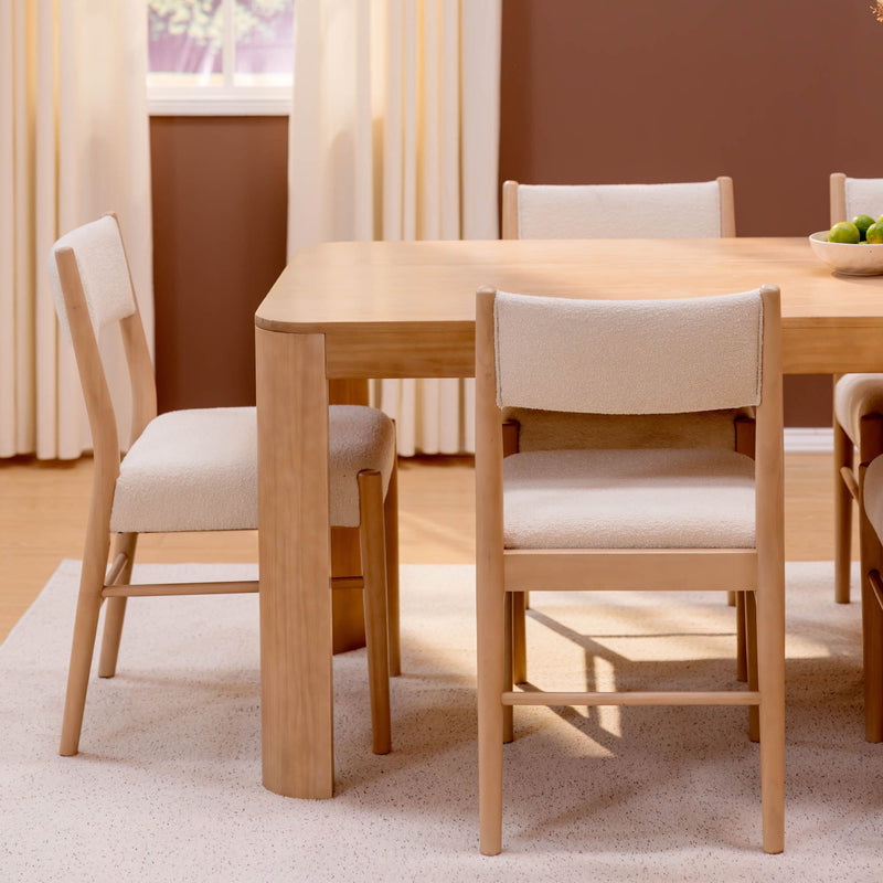 Light wood dining table with beige upholstered chairs in a modern minimalist neutral dining room with curtains and a bowl of green apples
