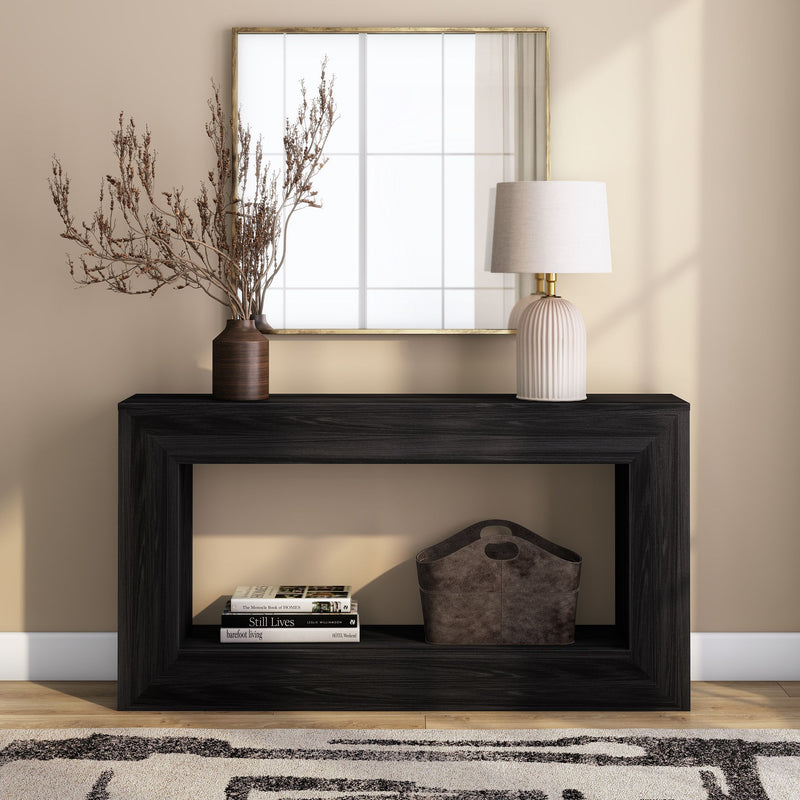 Minimalist modern entryway console table in black wood styled with a gold-framed mirror, ribbed ceramic table lamp, brown vase of dried branches, storage basket and stacked coffee-table books against a beige wall