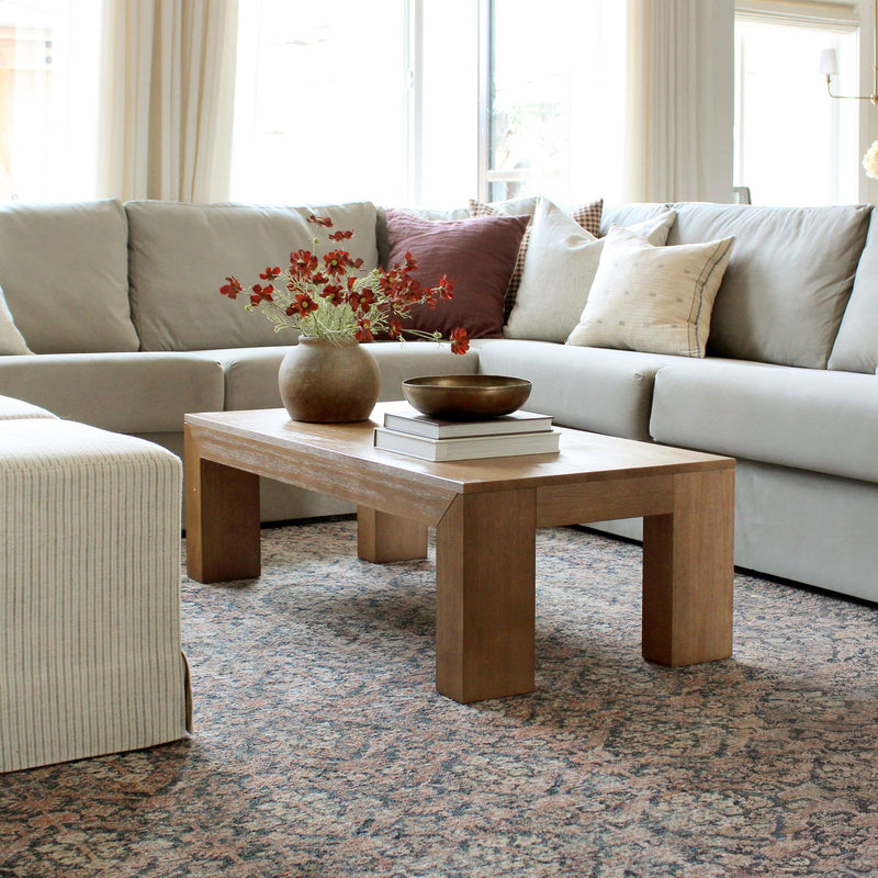 Light neutral living room with a light-gray sectional sofa, wooden coffee table topped with stacked books, bronze bowl and vase of red flowers, accent pillows and patterned area rug