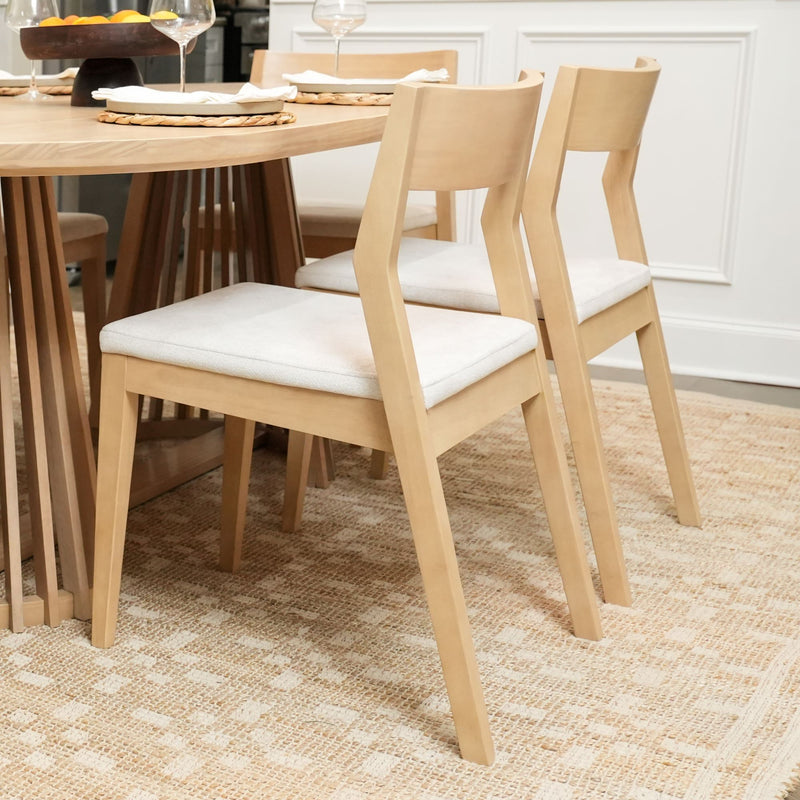 Modern wooden dining chairs with light fabric cushions around a round dining table on a patterned rug.