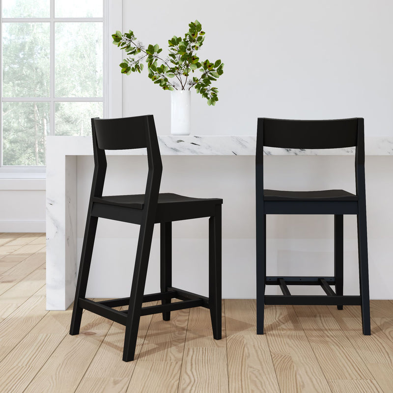 Modern kitchen interior with black bar stools at a marble counter, featuring a decorative vase with green foliage, natural lighting through a window, and wooden flooring.
