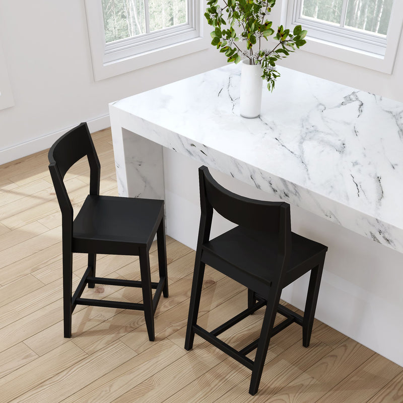 Two modern black bar stools next to a white marble table with a vase of green leaves, set on light wooden flooring by a window.