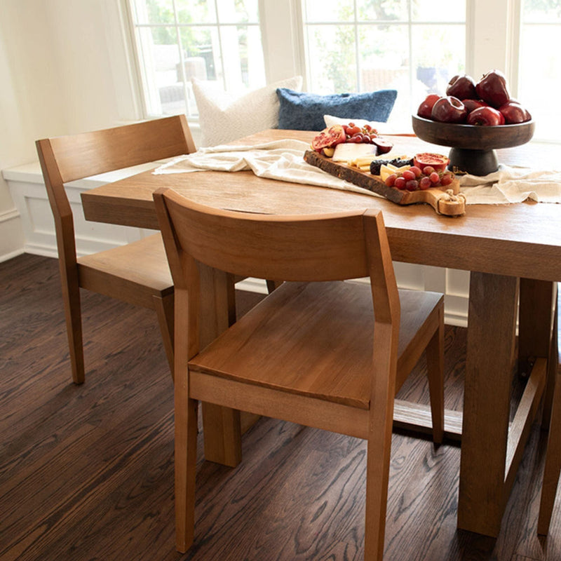Modern wooden dining table with two chairs, featuring a charcuterie board and fresh red apples, natural light from large windows.