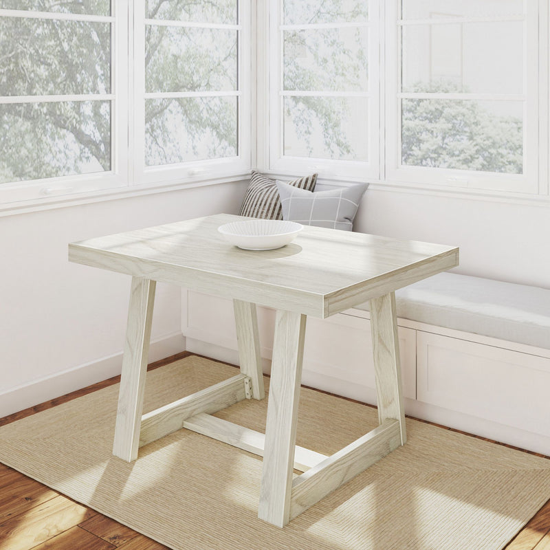 Sunlit modern farmhouse breakfast nook with whitewashed wooden table and ceramic bowl, window bench with striped and grid cushions, large white-framed windows, neutral rug and light hardwood floor
