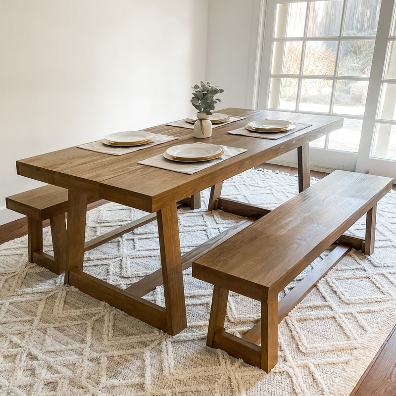 Rustic wooden dining table set with matching benches, place settings, and a vase on a textured cream rug near glass doors.