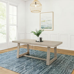 Light wood trestle dining table in modern farmhouse dining room with blue textured rug, rattan pendant light, large window and greenery centerpiece