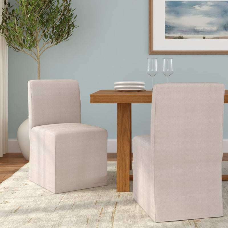 Neutral beige slipcovered dining chairs paired with a solid wood dining table, soft blue accent wall, area rug, potted plant and simple table settings in a modern minimalist dining room
