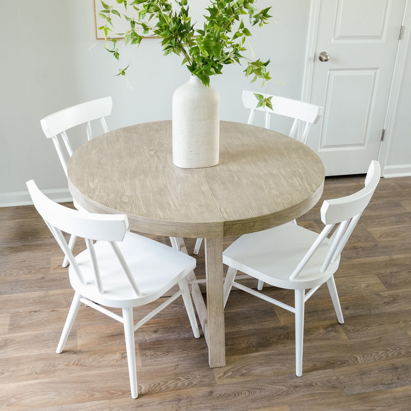 Round wooden dining table with white chairs and a vase of green leaves on wooden floor.