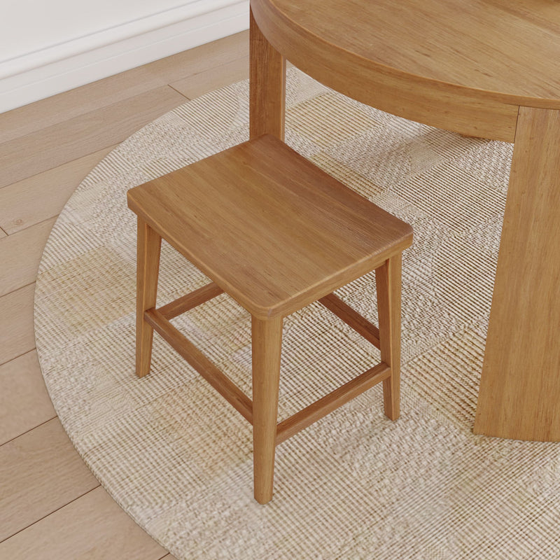 Wooden stool with rectangular seat next to a round wooden table on a woven rug in a bright room.