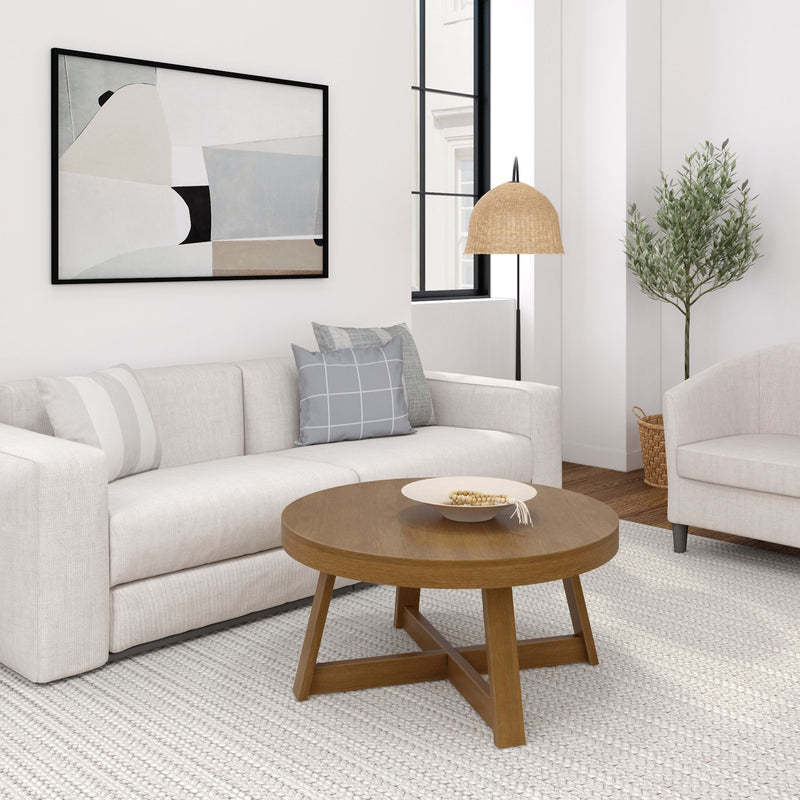 Modern neutral living room with beige upholstered sofa, gray grid throw pillows, round wooden coffee table on a textured ivory rug, abstract framed wall art, woven rattan pendant lamp and potted olive tree.