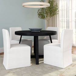 Modern round black dining table with four white slipcovered dining chairs in a bright minimalist dining room with pendant light, potted olive tree and light blue wall