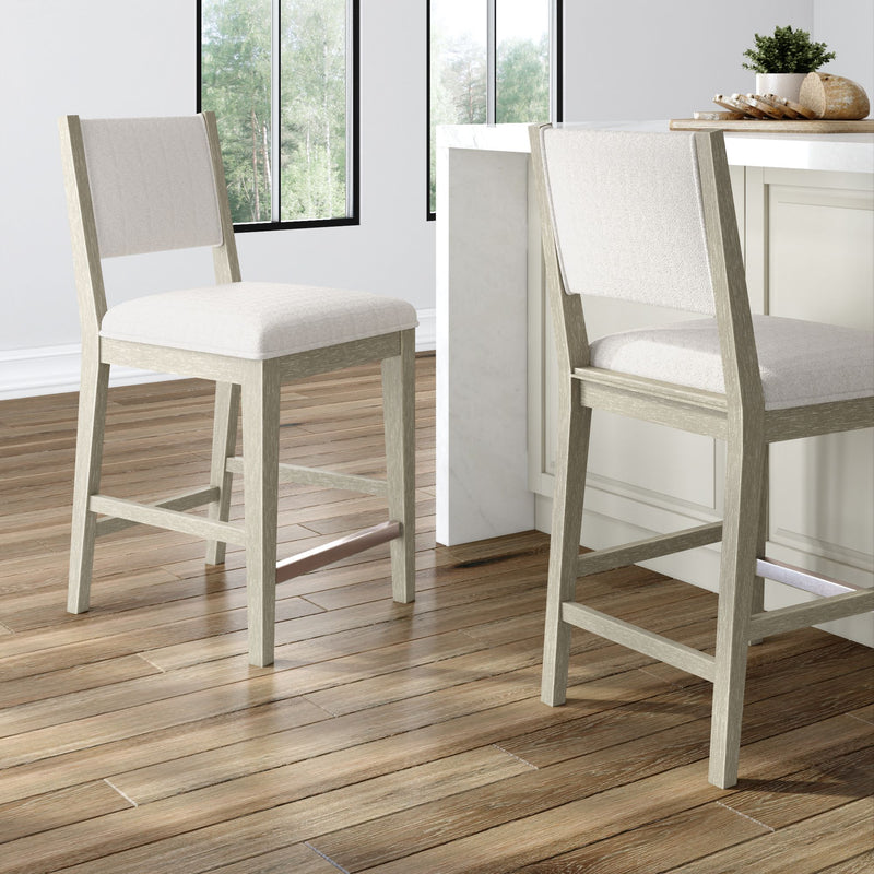 Two light-wood counter-height kitchen stools with beige upholstered seats at a white marble island on hardwood floors, modern farmhouse breakfast bar with large windows and natural light