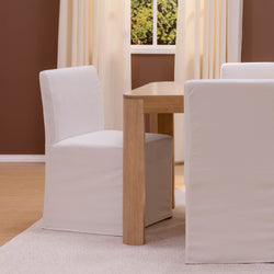White slipcovered dining chairs and light wood table in a minimalist neutral room with cream curtains and hardwood floor.