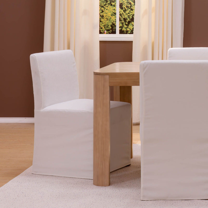 White slipcovered dining chairs and light wood table in a minimalist neutral room with cream curtains and hardwood floor.