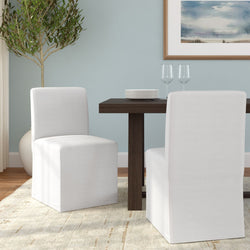 Modern dining room with white slipcovered chairs and dark wood table, stacked plates, wine glasses, light blue wall, potted olive tree and neutral area rug.