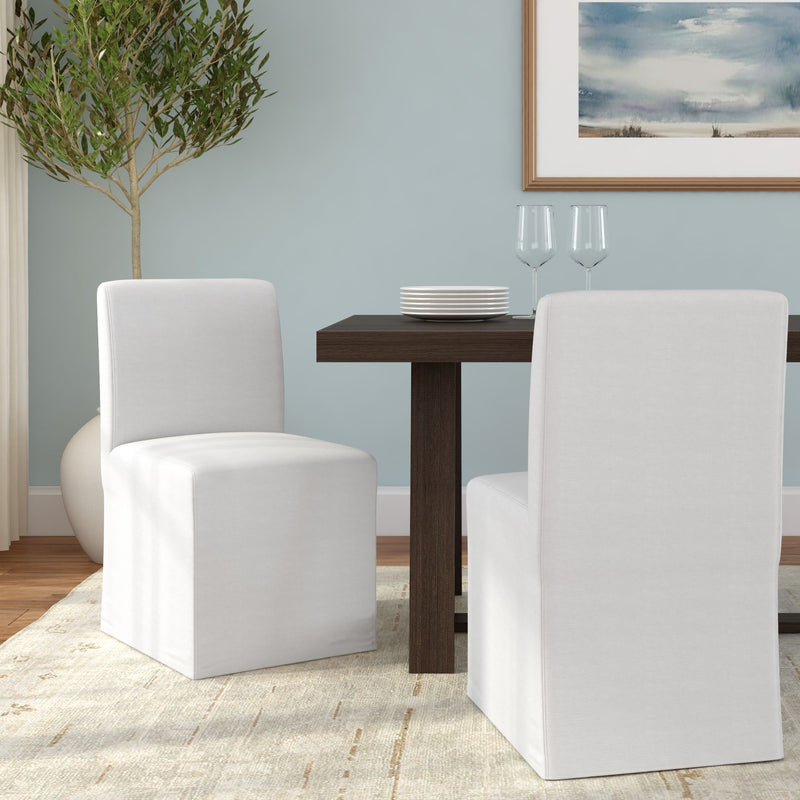 Modern dining room with white slipcovered chairs and dark wood table, stacked plates, wine glasses, light blue wall, potted olive tree and neutral area rug.