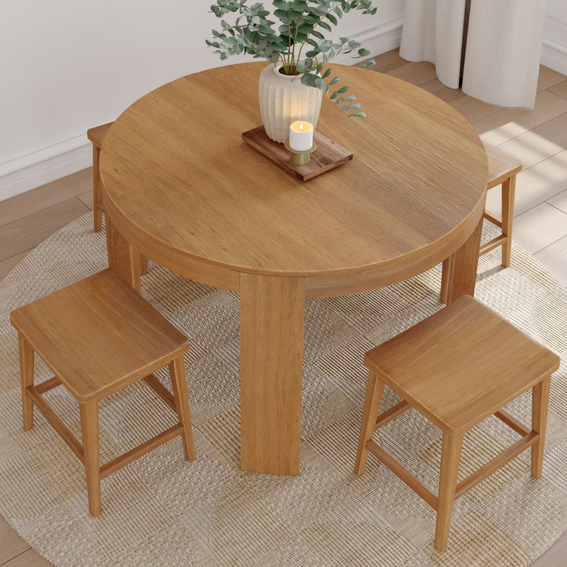 Round oak dining table with three matching wooden stools on a textured woven rug, styled with eucalyptus vase and candle in a minimalist sunlit dining nook