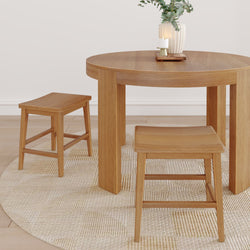 Round oak dining table with two matching wooden stools on a woven jute rug in a minimalist Scandinavian dining room styled with a ceramic vase of eucalyptus and a lit candle.