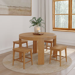 Round solid wood dining table with four matching stools on a round jute rug in a bright neutral dining room with eucalyptus vase centerpiece and natural light from window