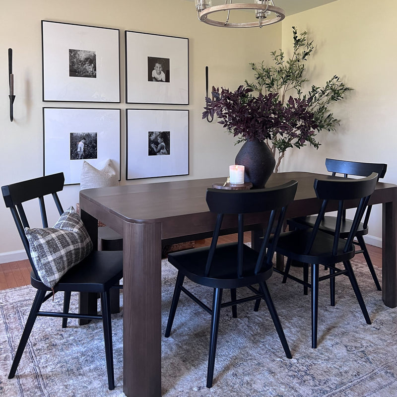 Elegant dining room with a dark wood table, black chairs, a vase with purple foliage, framed black-and-white photos, and a lit candle on a stylish rug.