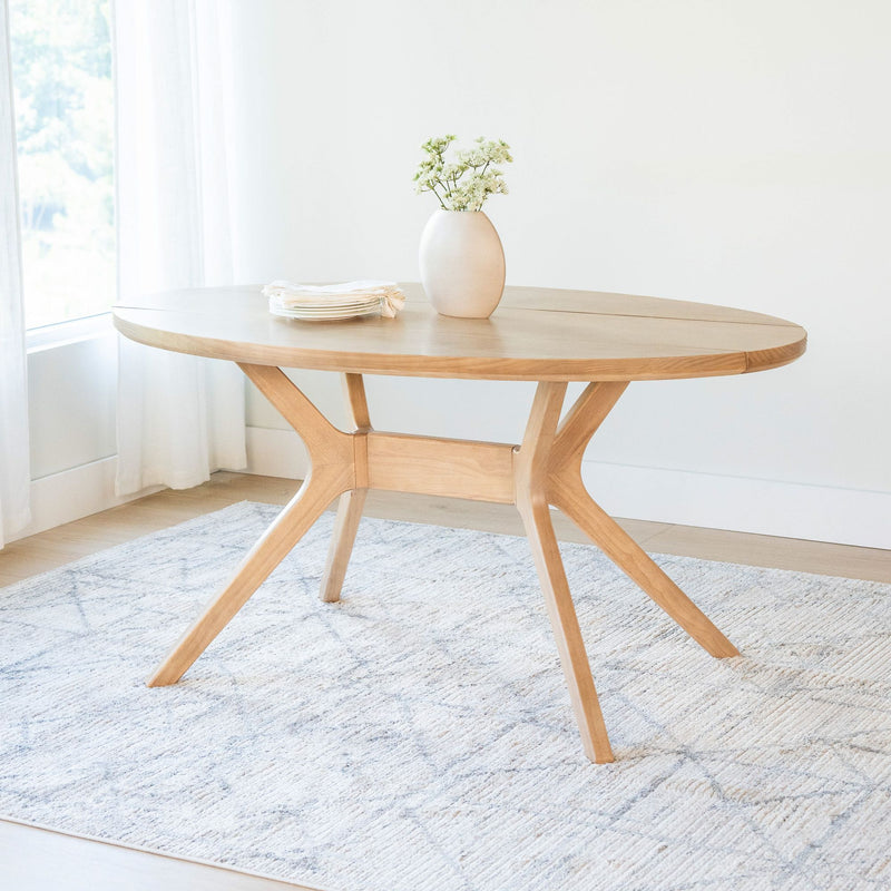 Oval wooden dining table with modern design, featuring a centerpiece vase and neatly stacked plates, placed on a light-patterned rug in a bright room.