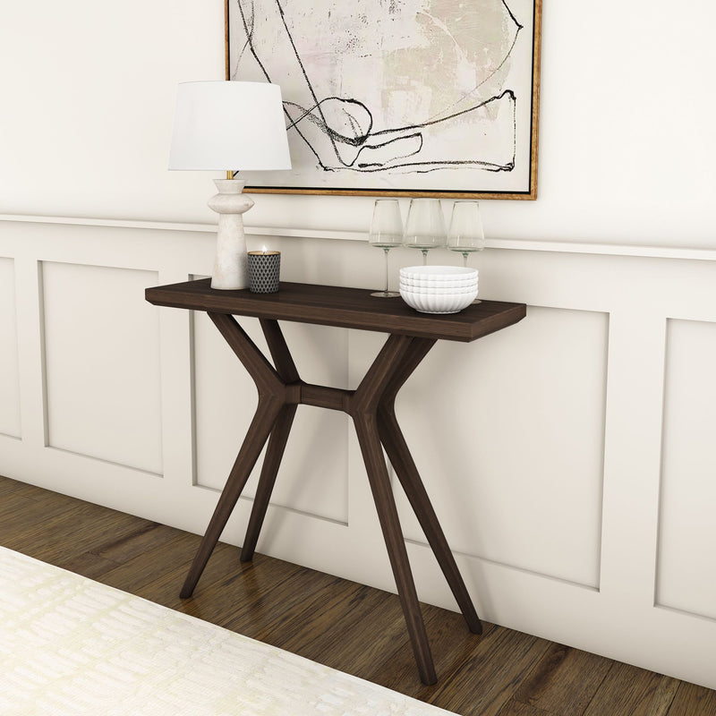 Modern dark wood console table with sculptural legs in a white-paneled entryway styled with a table lamp, candle, glassware, white bowls, and abstract wall art.