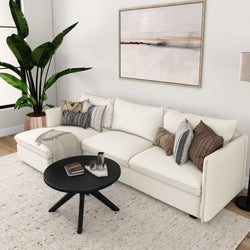 Modern living room with cream sectional sofa, decorative cushions, large houseplant, abstract artwork, and black coffee table on textured rug.