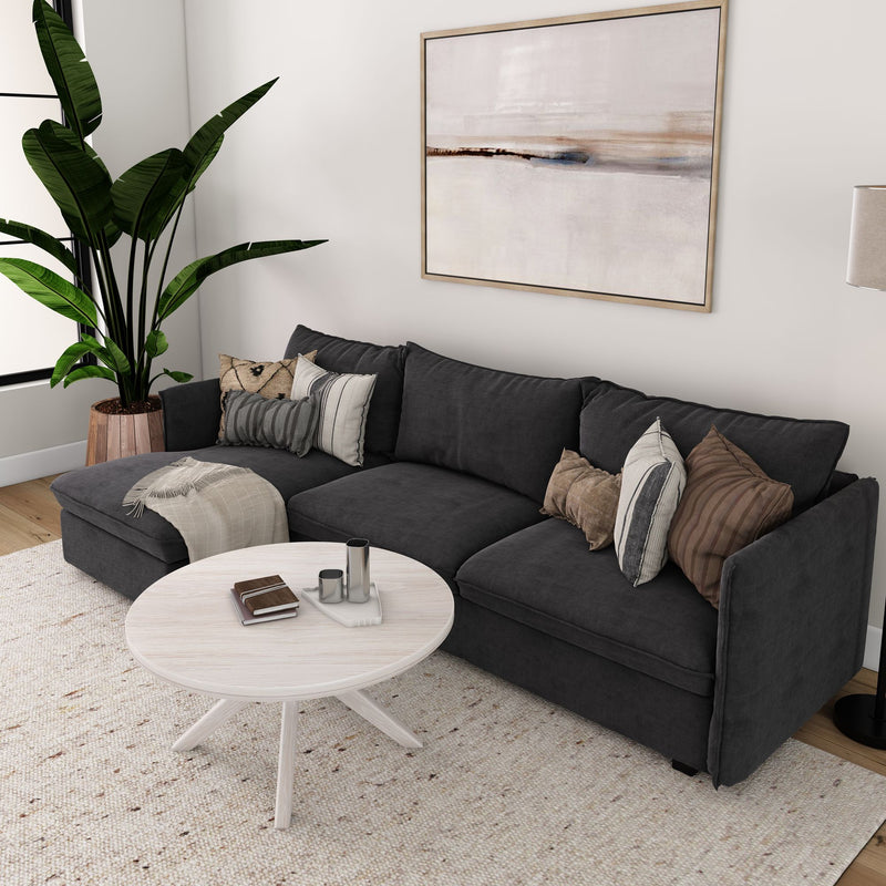 Modern living room with dark grey sectional sofa, decorative pillows, large indoor plant, abstract wall art, and round coffee table.