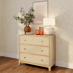 Natural wood three-drawer rattan dresser styled with a ceramic table lamp, terracotta vase of greenery and amber glass candles on hardwood floor against a textured plaster wall, boho coastal bedroom decor