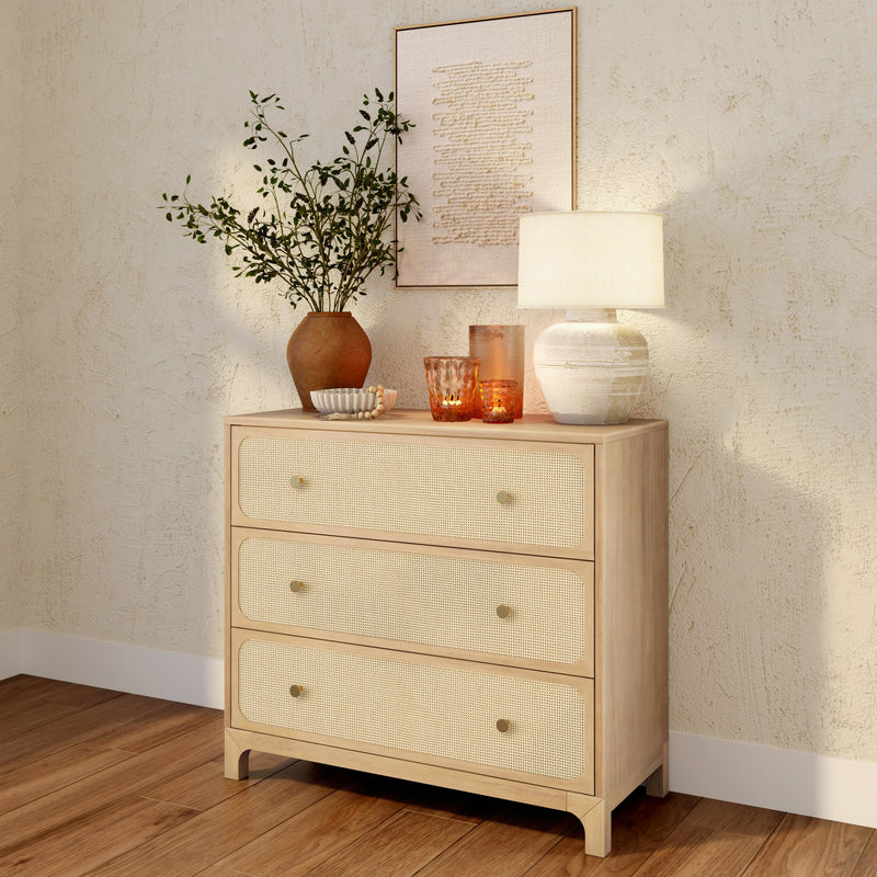 Natural wood three-drawer rattan dresser styled with a ceramic table lamp, terracotta vase of greenery and amber glass candles on hardwood floor against a textured plaster wall, boho coastal bedroom decor