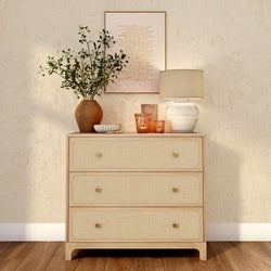 Rattan-front three-drawer wooden dresser in neutral boho decor with ceramic table lamp, terracotta vase of greenery, glass candleholders and textured wall art on hardwood floor