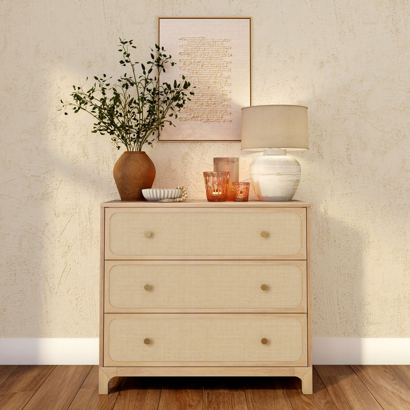 Rattan-front three-drawer wooden dresser in neutral boho decor with ceramic table lamp, terracotta vase of greenery, glass candleholders and textured wall art on hardwood floor