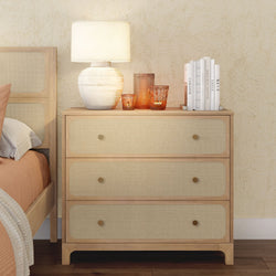 Rattan-front wooden three-drawer bedside dresser with ceramic table lamp, amber glass candles and stacked books in a neutral boho bedroom