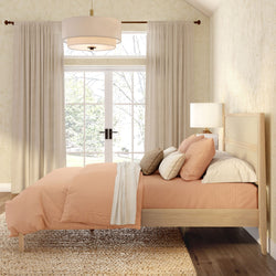 Cozy modern boho bedroom with peach duvet on natural wood bed and rattan headboard, layered pillows, bedside lamp and pendant light, jute rug, French doors with beige curtains