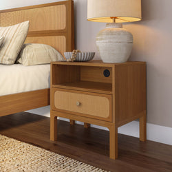 Wooden bedside table with woven rattan drawer, ceramic table lamp and neutral pillows against a cane headboard in a cozy bedroom.