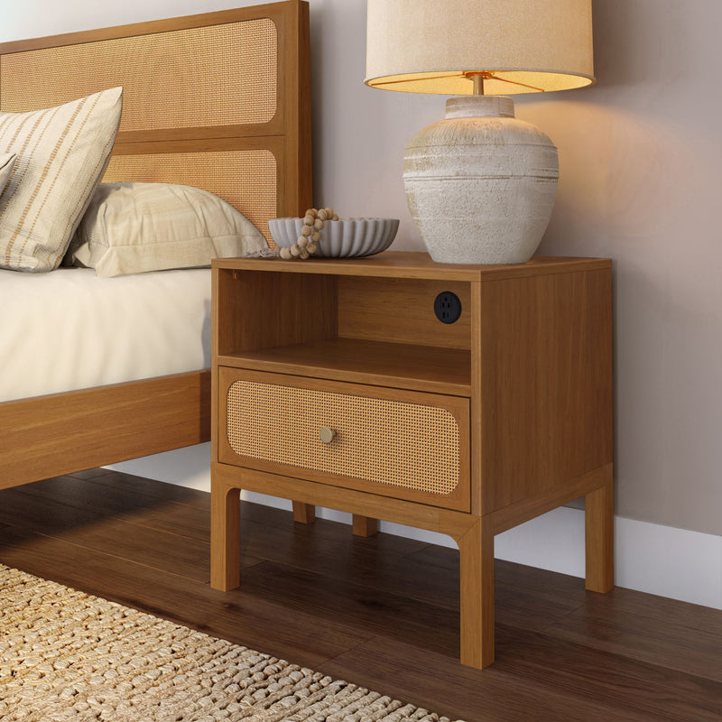 Wooden bedside table with woven rattan drawer, ceramic table lamp and neutral pillows against a cane headboard in a cozy bedroom.