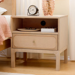 Light wood nightstand with cane-front drawer, black round alarm clock, pink bubble glass vase and reading glasses — modern bedroom decor