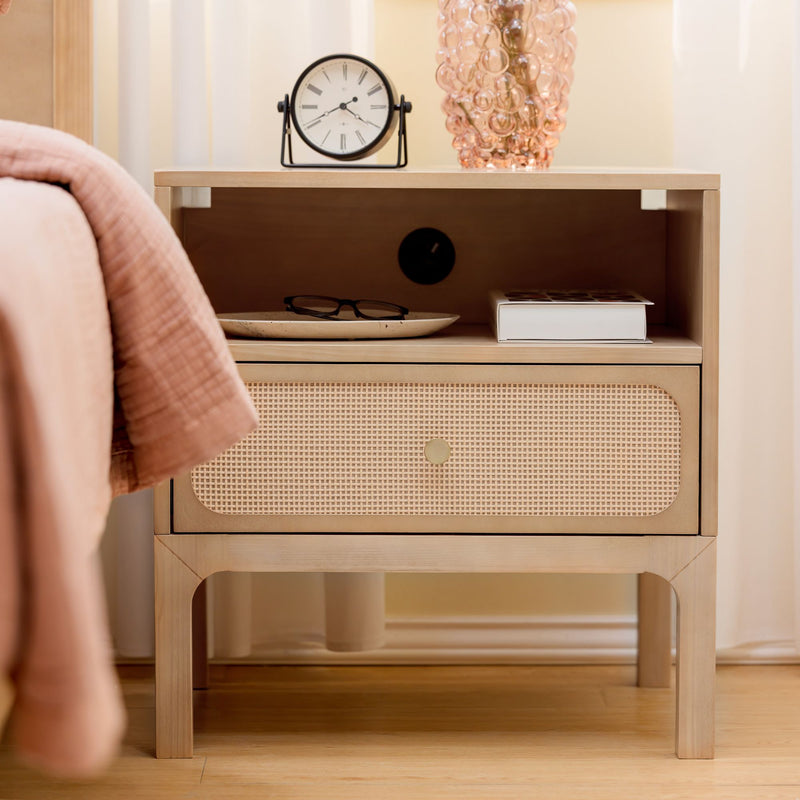 Light wood Scandinavian rattan bedside table nightstand with cane drawer, alarm clock, pink glass vase, book and reading glasses beside pink bedding — minimalist bedroom decor