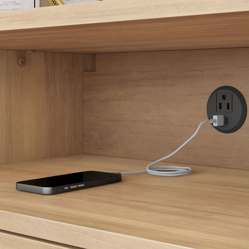 Black smartphone charging on a wooden shelf inside a cabinet, USB charging cable plugged into a round wall outlet