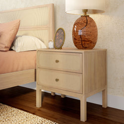 Boho-style wooden two-drawer nightstand with woven rattan fronts, amber glass table lamp, small round mirror and candle next to a beige bed in a neutral bedroom décor