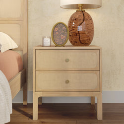 Natural wood bedside table with woven cane drawer fronts (rattan nightstand), styled with amber glass lamp, scented candle and round floral frame in a boho bedroom