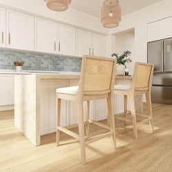 Modern kitchen interior with light wood flooring, white cabinets, marble island, and woven chairs.