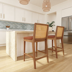 Modern coastal kitchen with marble waterfall island, light hardwood floors, white shaker cabinets, blue subway tile backsplash, wooden rattan-backed bar stools, wicker pendant lights and stainless steel refrigerator