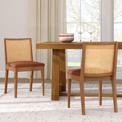 Wooden dining table with two cane-back dining chairs with rust-colored cushions, ceramic bowls and wine glasses on table in a bright neutral dining room by a large window
