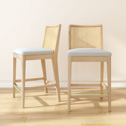 Pair of counter-height light wood bar stools with rattan cane backs and beige upholstered seats on hardwood floor — modern Scandinavian kitchen island seating