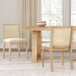 Light wood dining table with rattan cane-back chairs and upholstered white seats, ceramic bowls and wine glasses in a bright neutral dining room